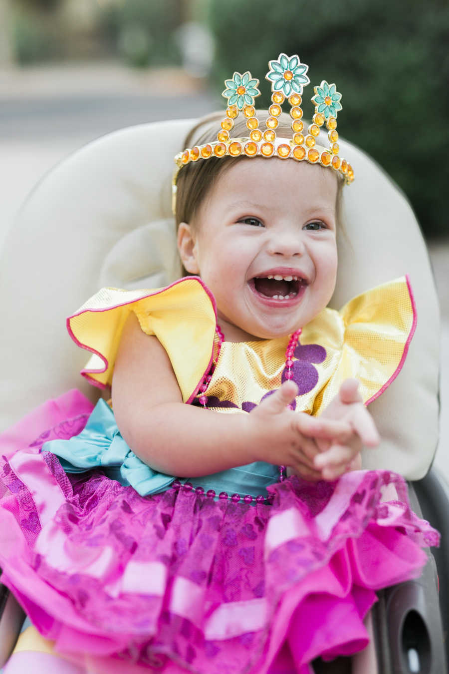 Baby with down syndrome and heart defect sits smiling wearing colorful dress and crown