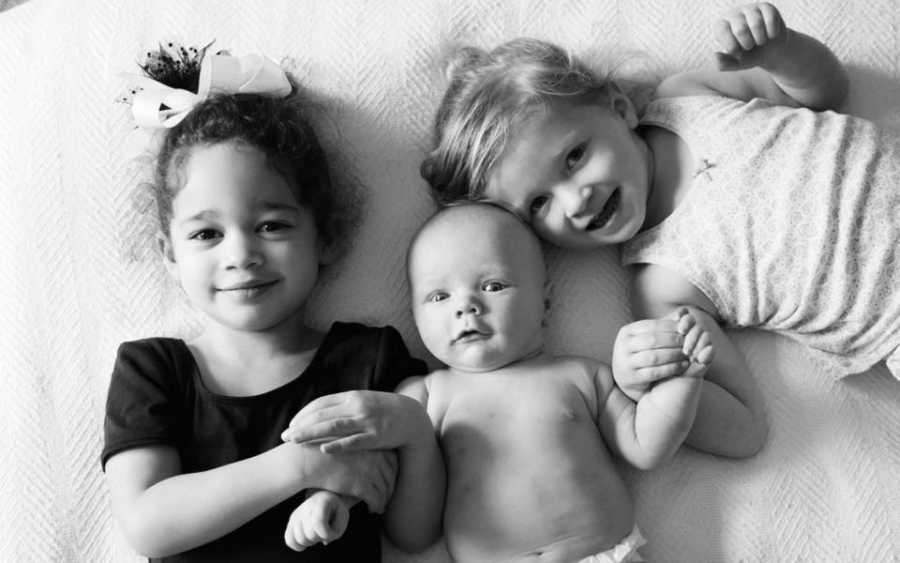 Sisters lay on their back smiling with their baby brother laying between them