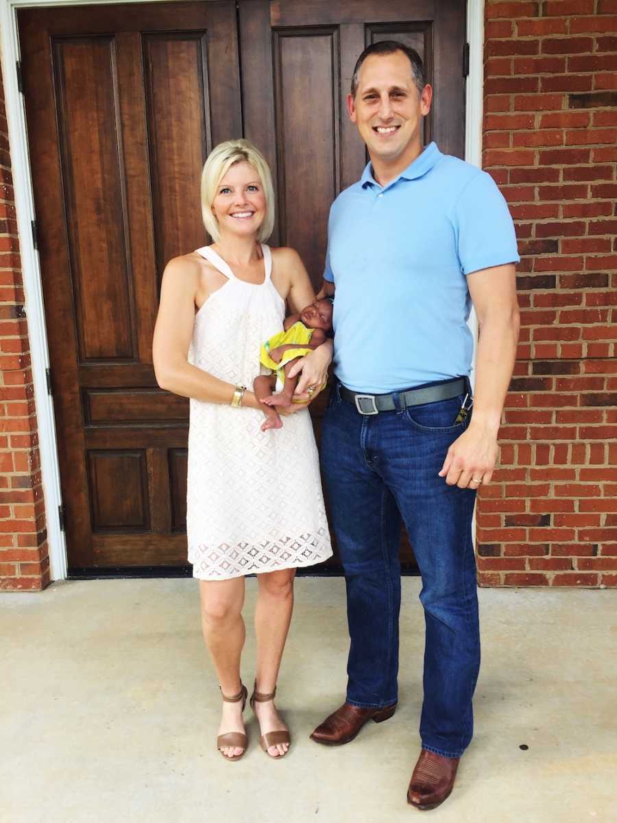 Husband and wife stand outside of brick building while wife holds adopted newborn