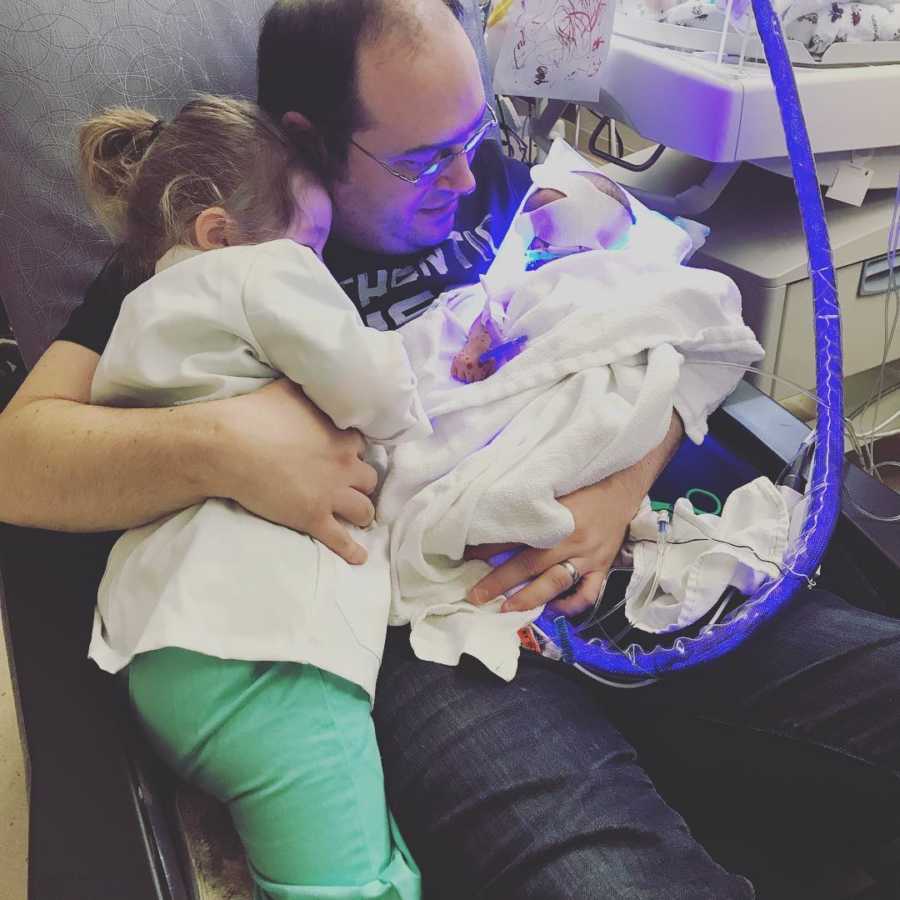 Father sits in chair in NICU holding newborn in one arm and older daughter in the other