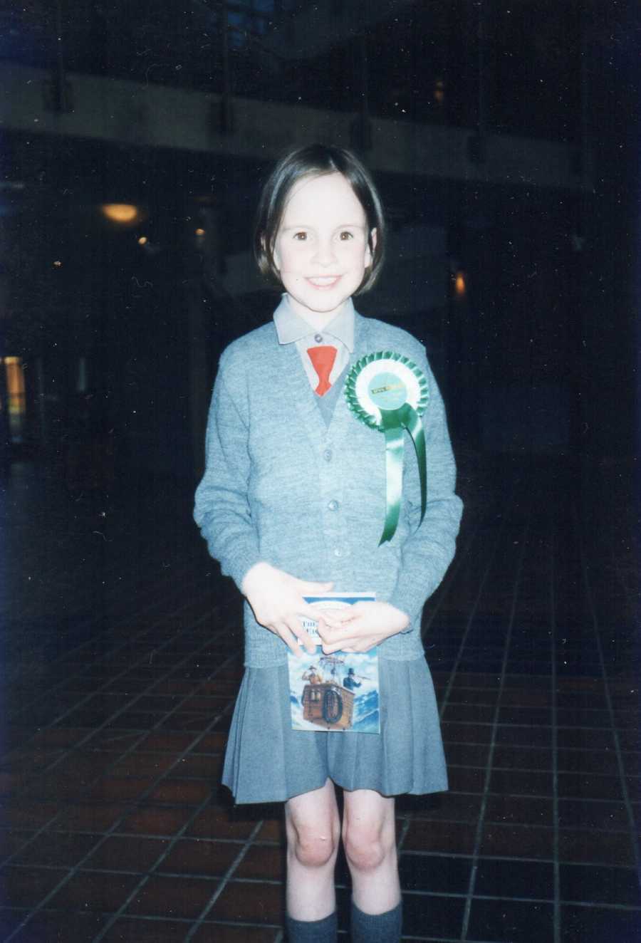 Young girl with Exomphalos stands in school uniform with green and white ribbon on her chest