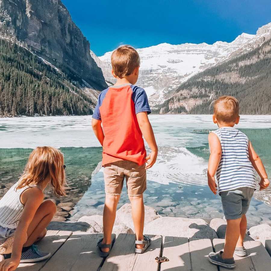 Three young siblings stand on dock looking out at body of water in between mountains