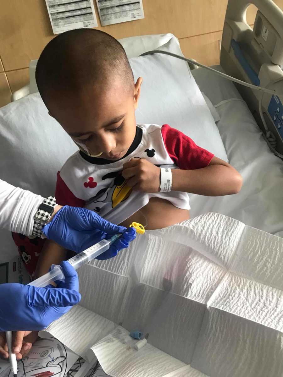 Little boy with cancer holds up shirt as nurse holds needle