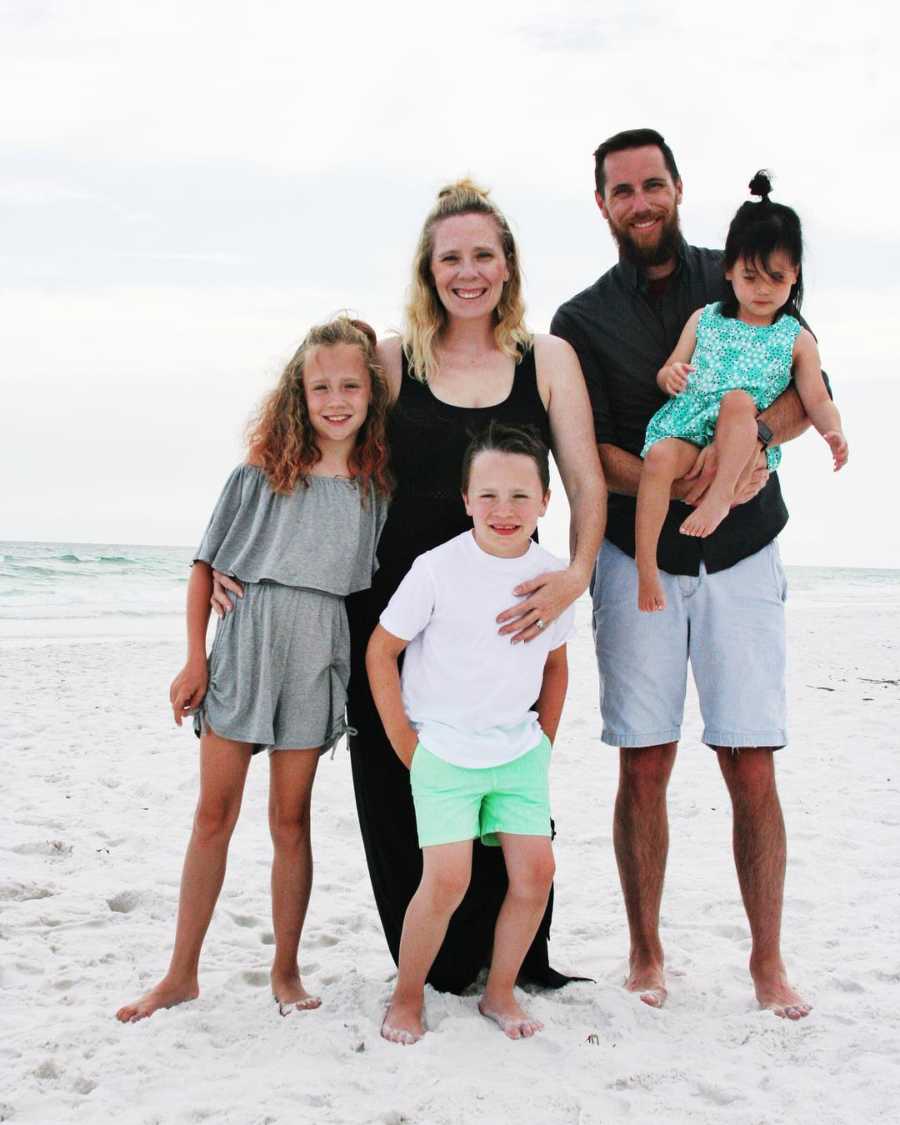 Husband and wife stand on beach with their two biological kids while husband holds adopted kid