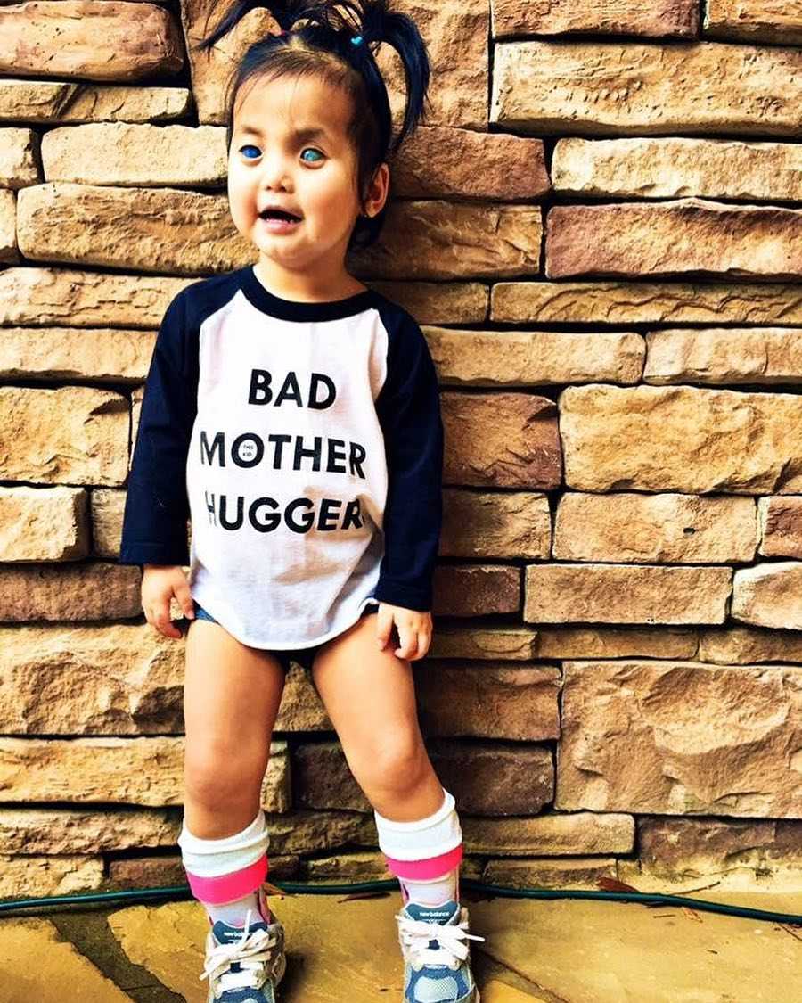 Blind and deaf little girl stands smiling against stone wall wearing shirt that says, "bad mother hugger"