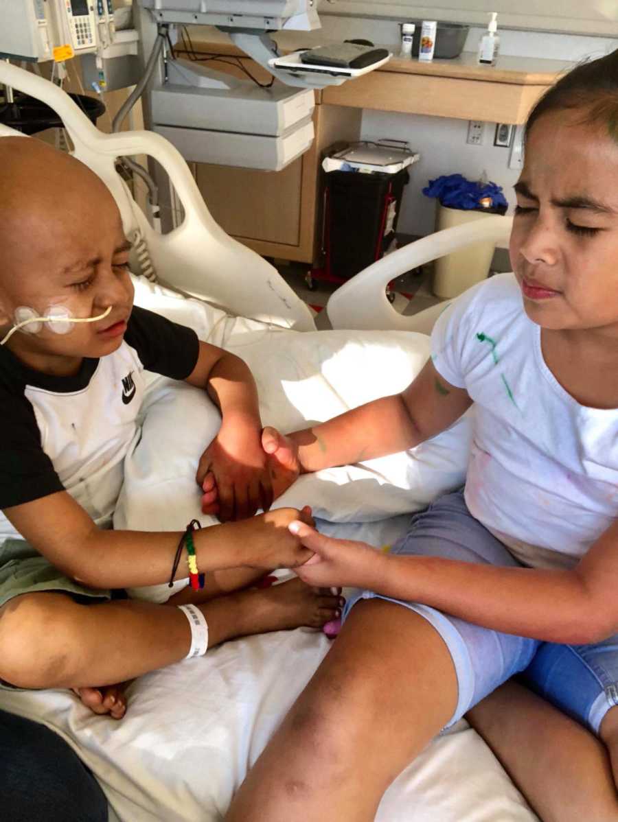 Little boy with cancer sits in hospital bed holding hands with sister as they pray