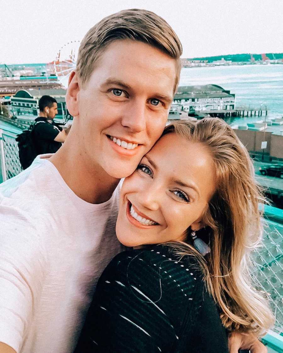 Husband and wife stand smiling with pier and ocean in background