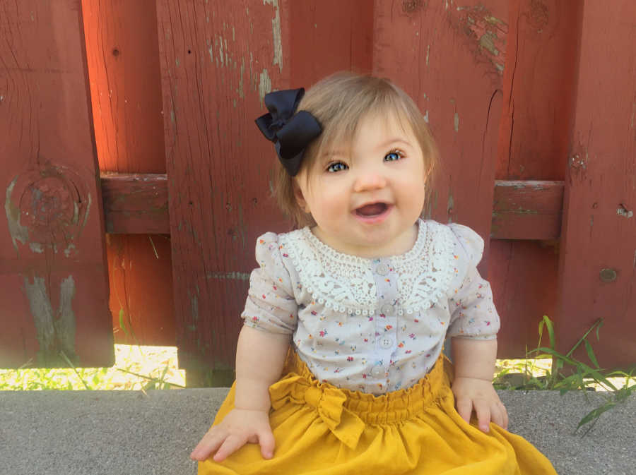 Little girl with down syndrome sits on side walk with red fence behind her