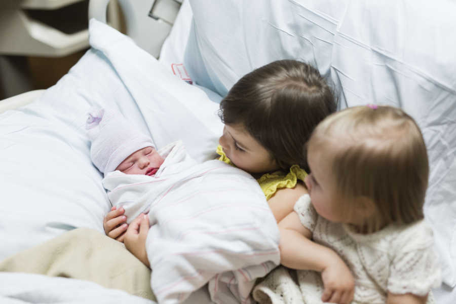 Two little girls sit in hospital bed holding their newborn sibling