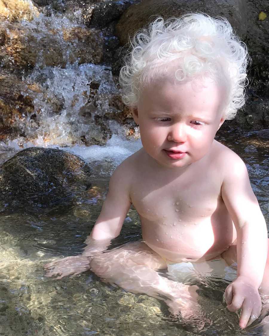 Albino baby sits in shallow water by small waterfall