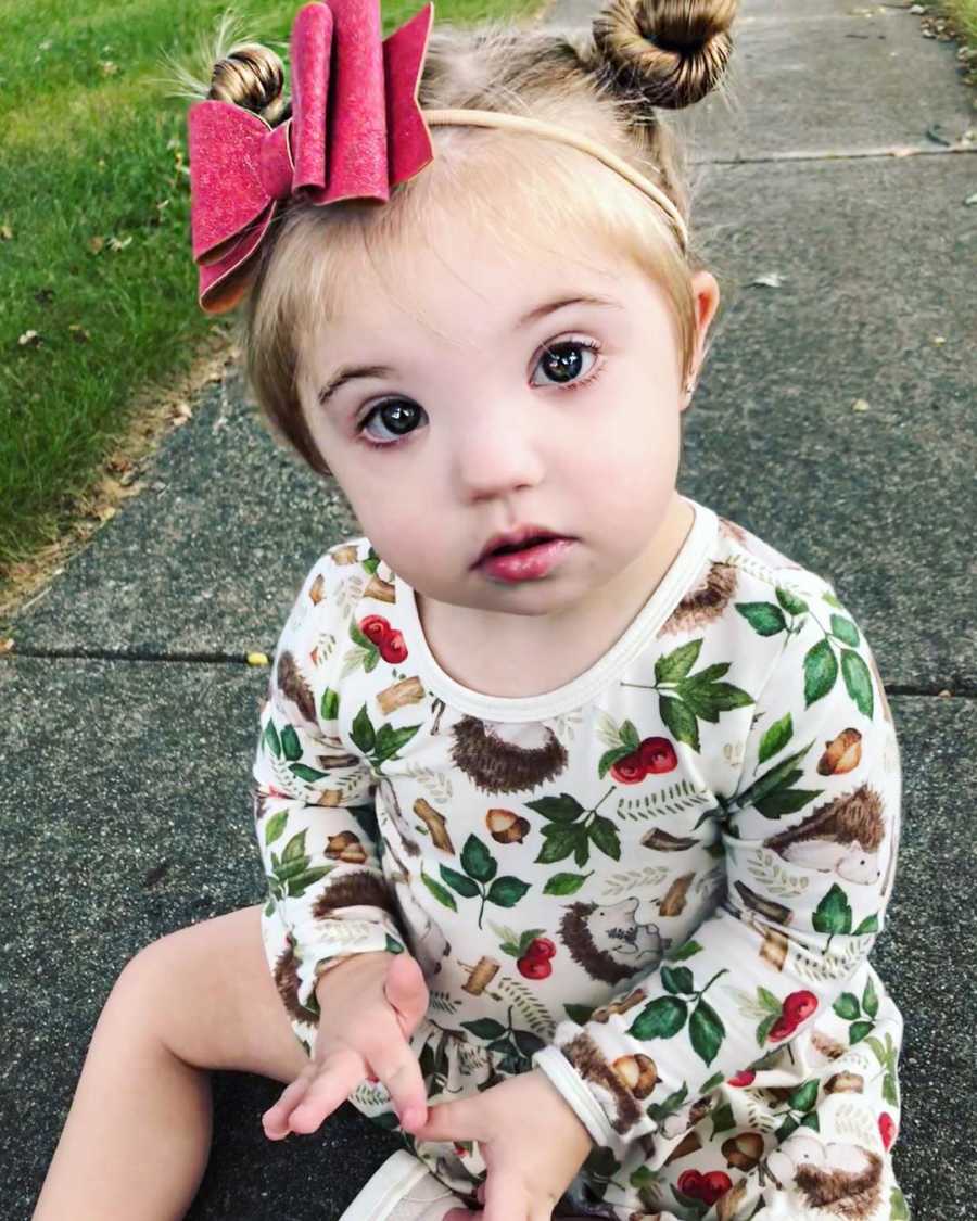 Little girl with down syndrome sits on sidewalk with big pink bow in her hair