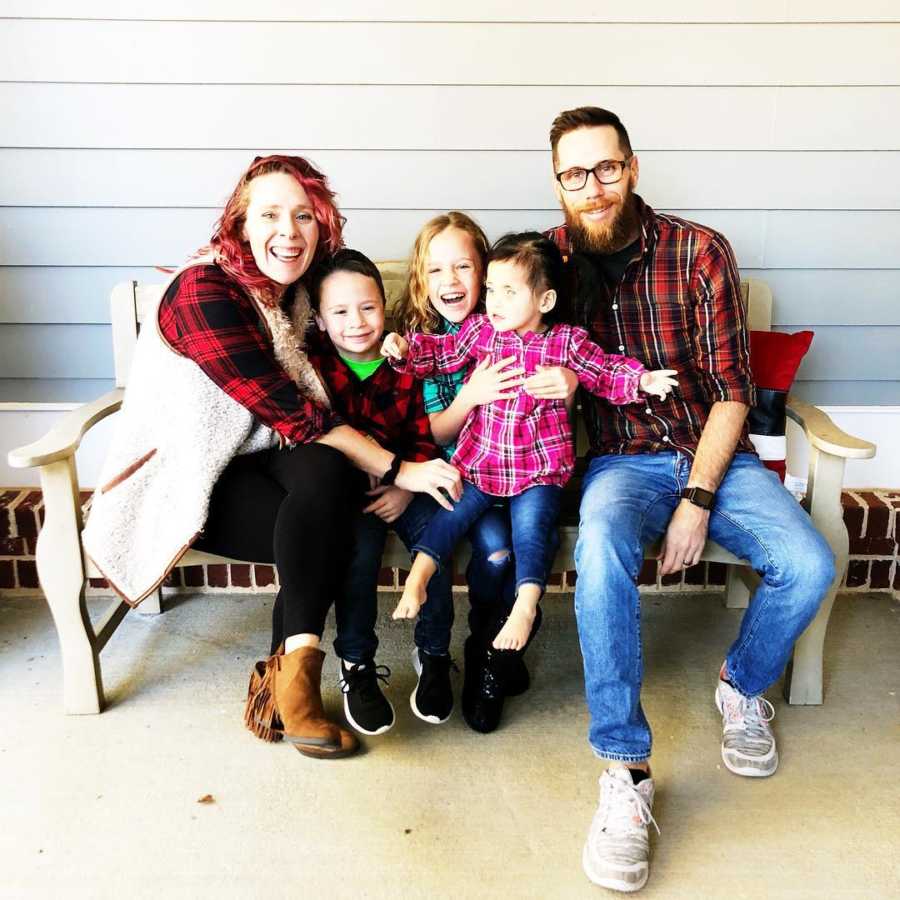 Husband and wife sit on bench outside home with their three children in between them