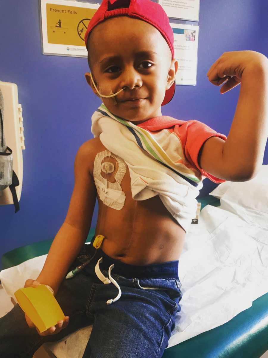 Little boy with cancer sits in doctor's office holding shirt up to show bandage