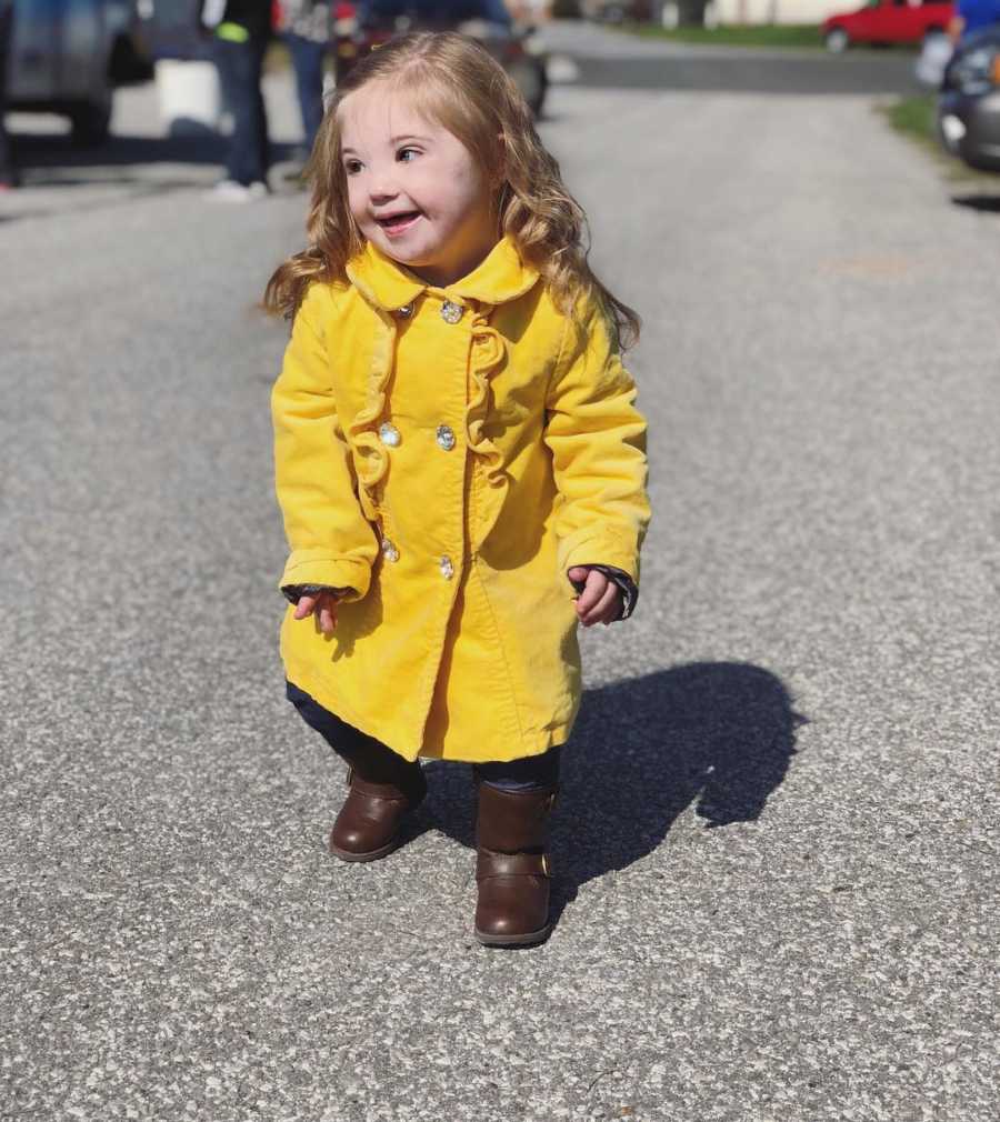 Little girl with down syndrome stands smiling in street wearing long yellow coat