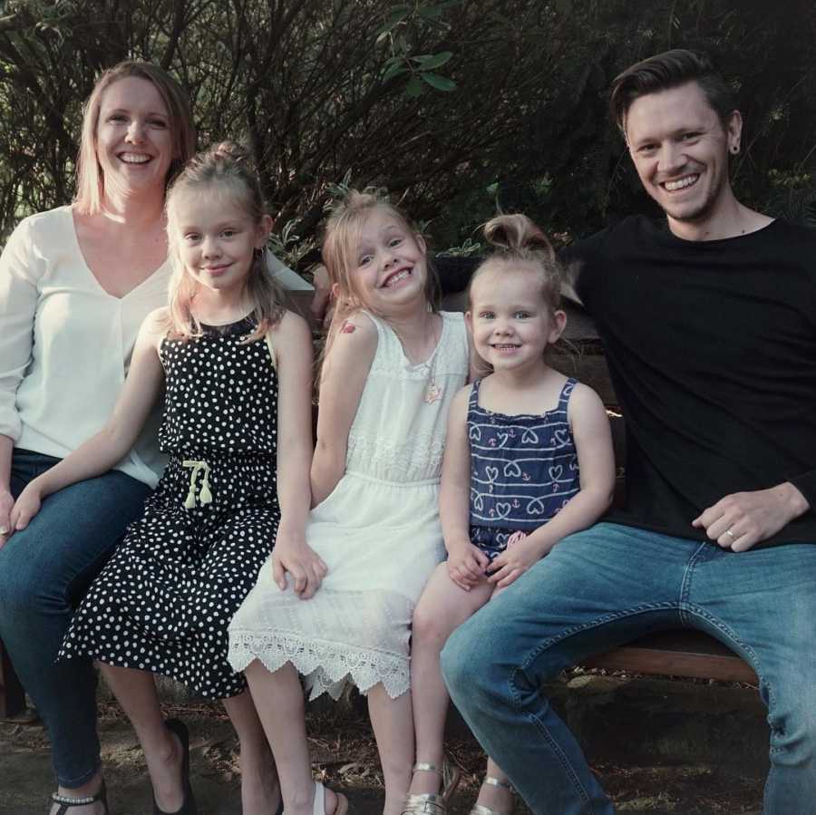 Young girl sits on bench outside with two younger sisters, mom, and dad