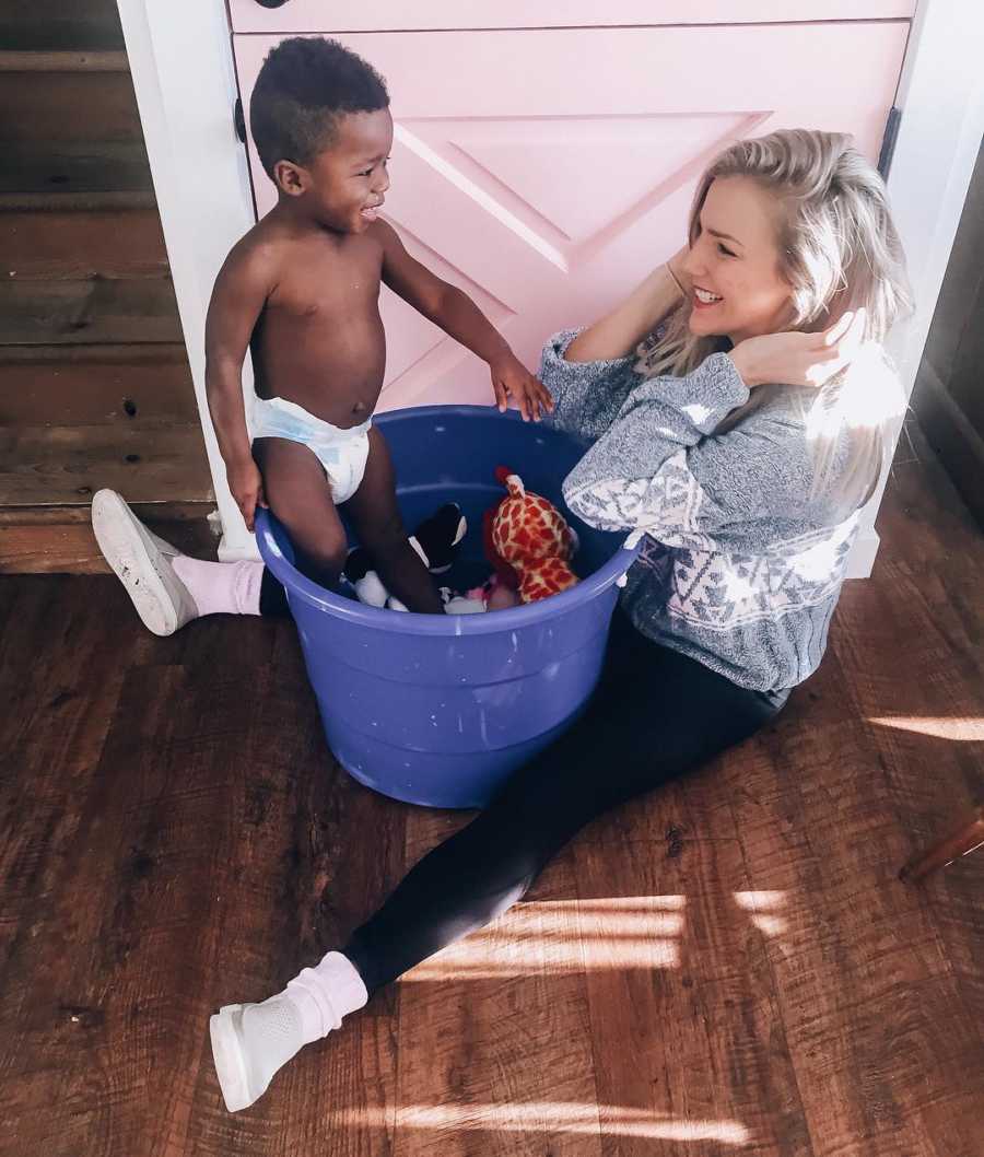 Mother smiles as she looks at baby boy who is standing in blue bin in-between her legs