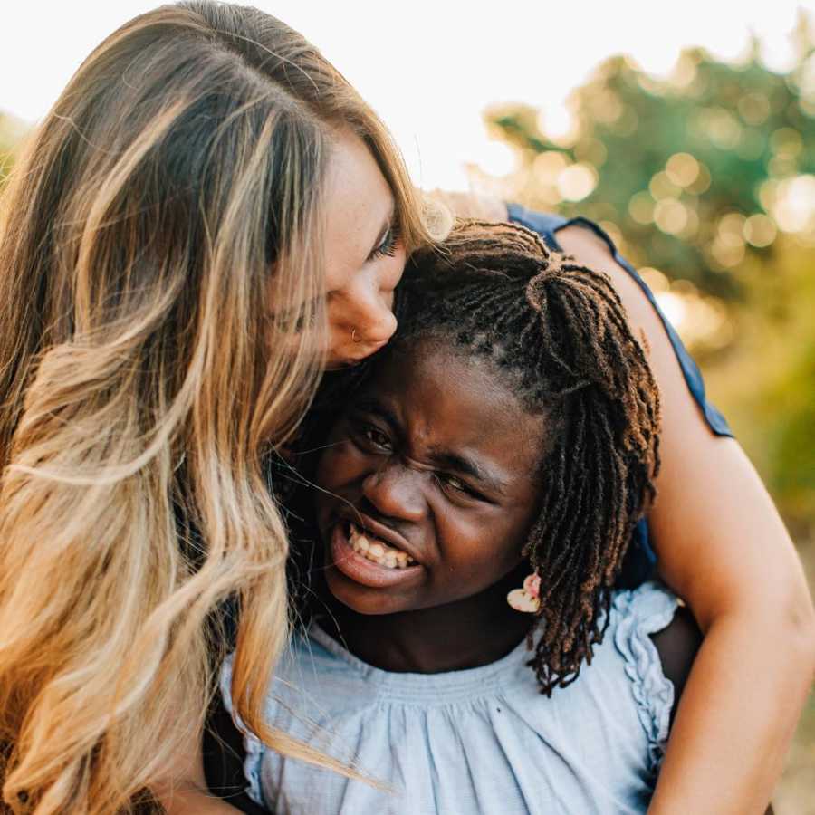 Mother stands behind adopted daughter kissing her forehead as daughter smiles