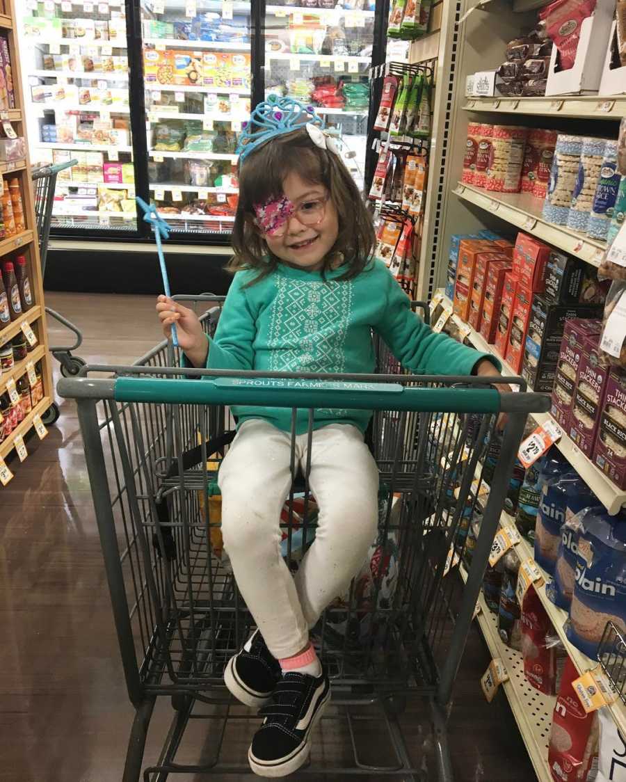 Little girl with strabismus sits smiling in shopping cart in aisle of grocery store
