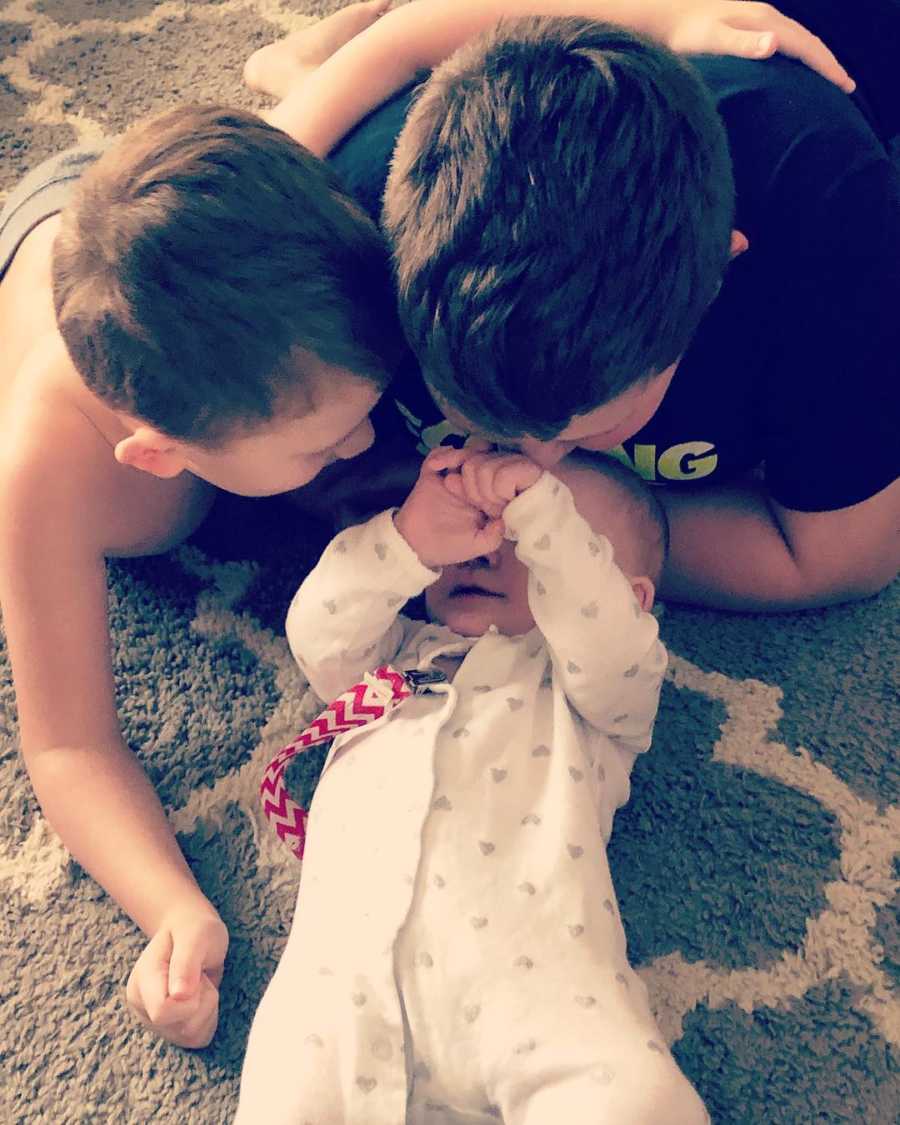 Baby lays on her back on floor while her older brothers lay beside her looking at her