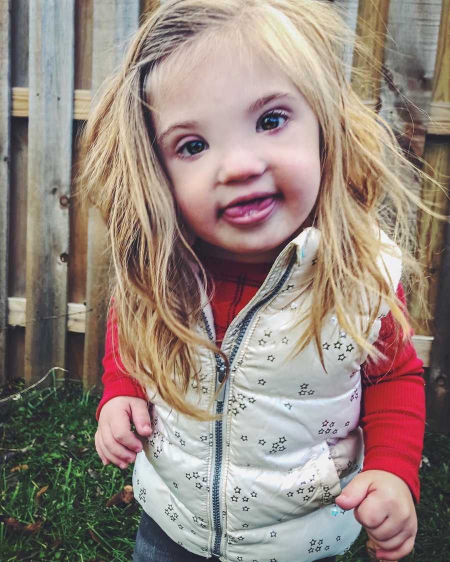 Little girl with down syndrome stands smiling in yard with wood fence in background