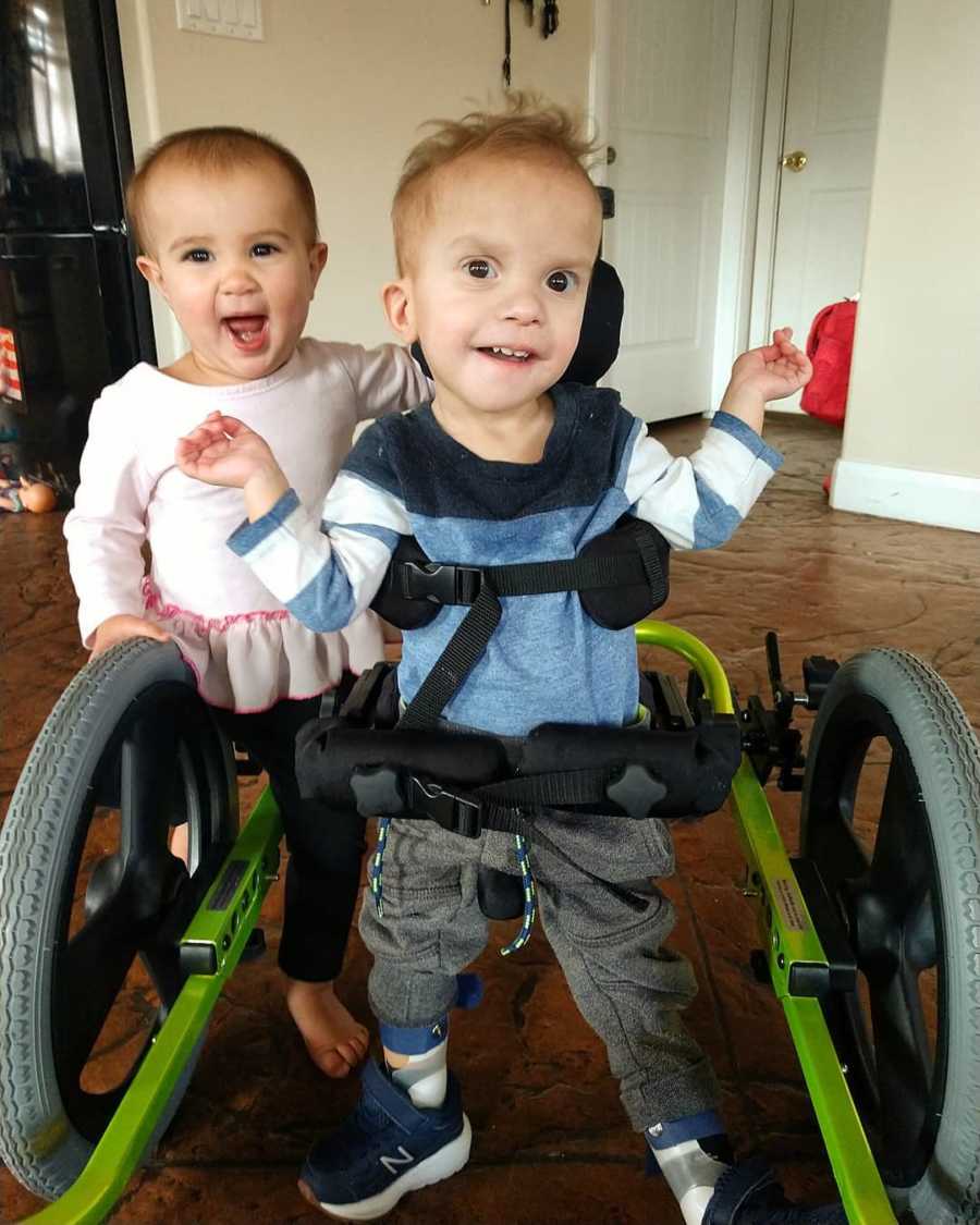Baby with unknown disease stands smiling strapped to walker with baby girl behind him
