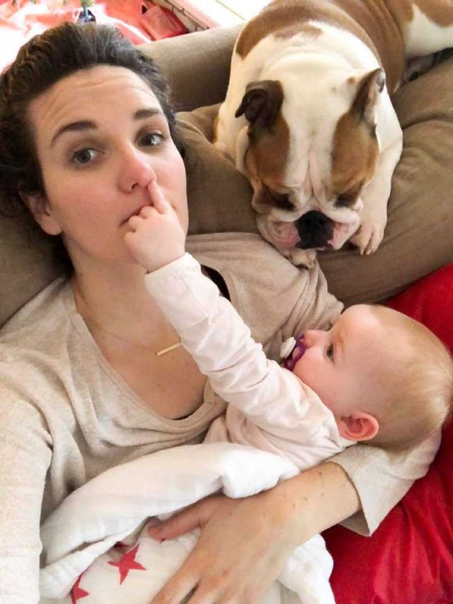 Mother sits on couch with dog laying by her shoulder and baby in her arms putting finger up her nose