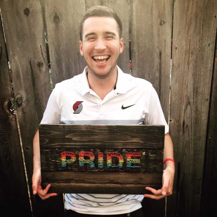 Gay man stands smiling in front of wooden fence holding sign that says, "pride" in rainbow letters