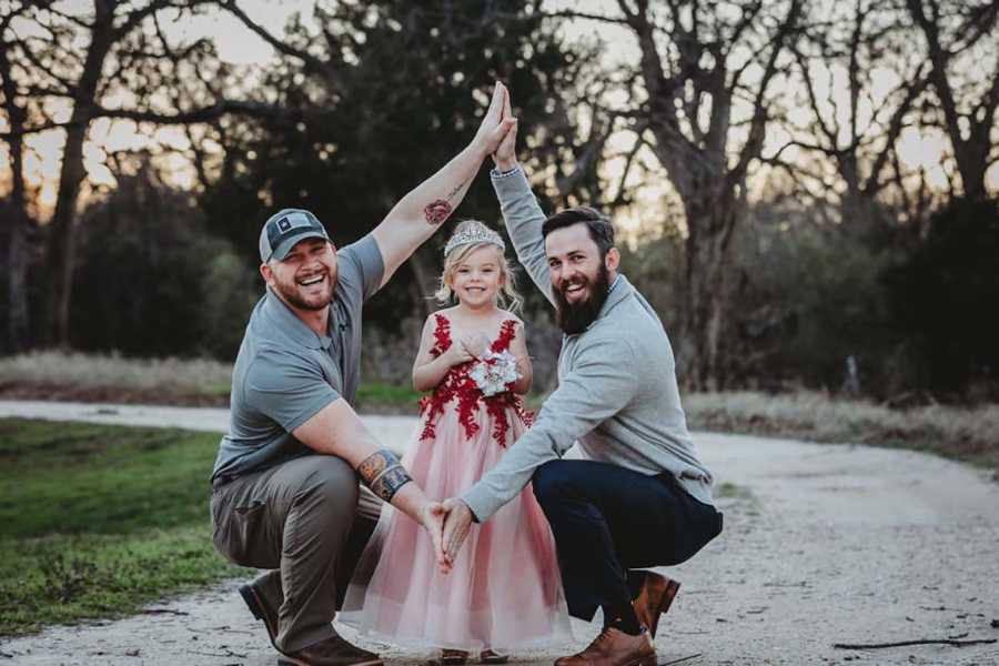 Little girl stands outside wearing dress and crown as her dads kneel on either side of her with hands holding