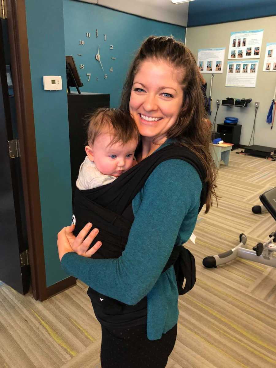 Woman who works as chiropractic assistant stands smiling in office with her baby swaddled to her