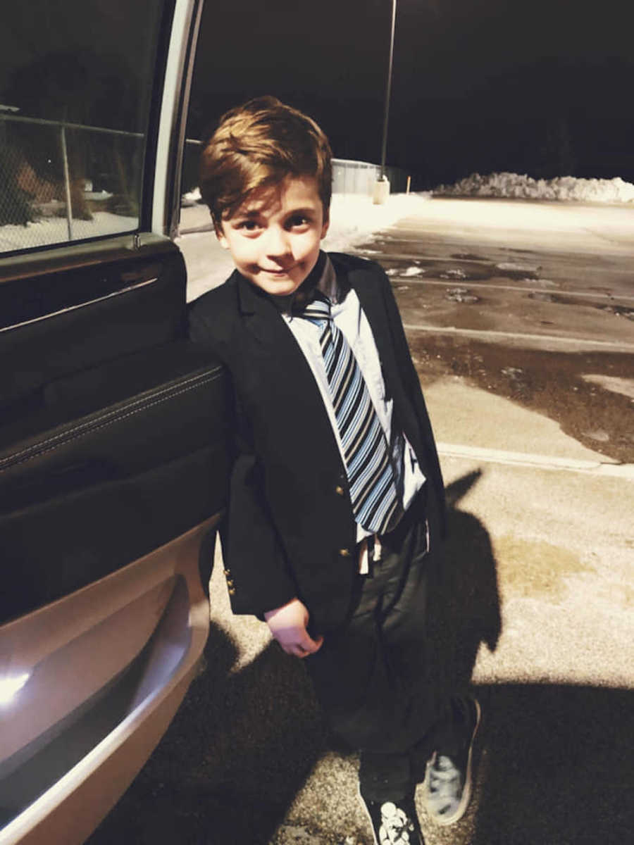 Little boy who doesn't listen stands smiling in suit and tie beside open car door