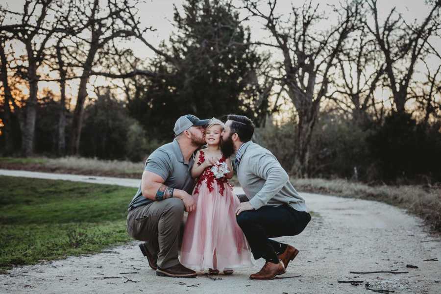 Young girl stands outside in dress and crown as her two dads kiss her cheeks