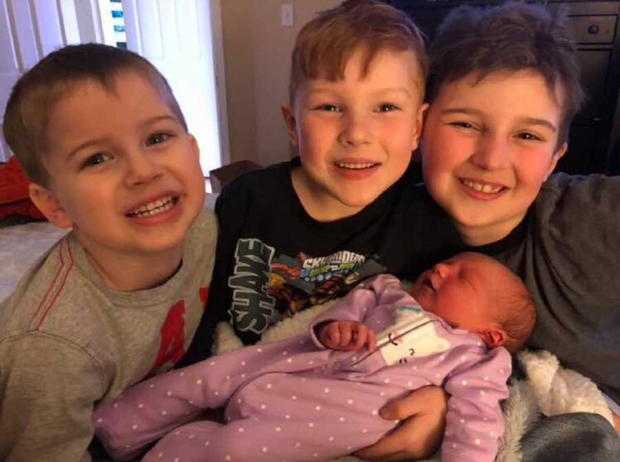Three young boys smiles as they hold baby sister
