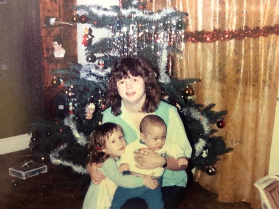 Mother crouches down in front of Christmas tree with two young kids in her lap