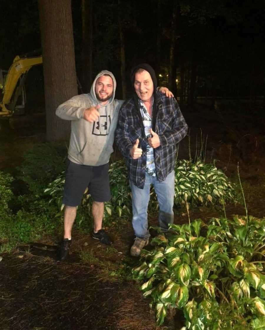Man who has brain tumor stands outside in garden holding thumbs up beside son