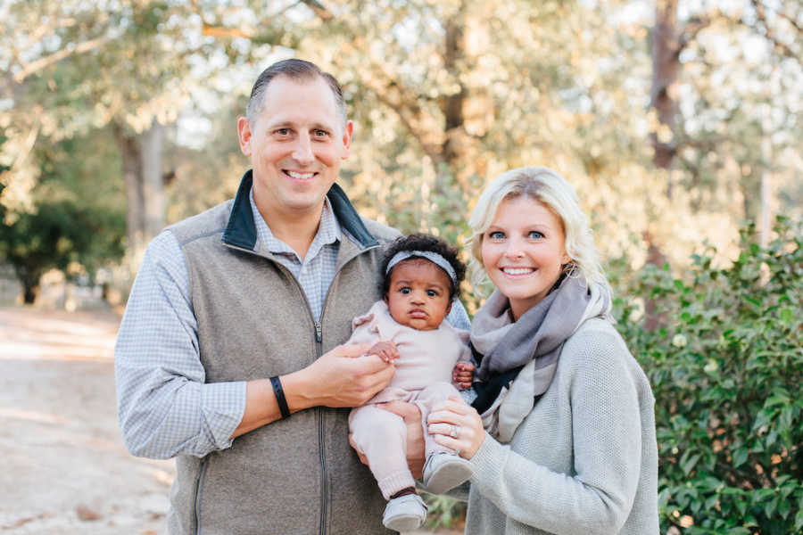 Husband and wife stand outside smiling as they hold their adopted daughter