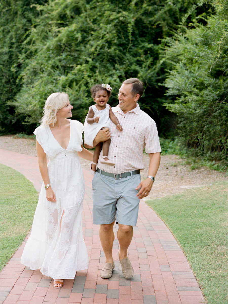 Father smiles at his adopted daughter he holds her while walking along path beside wife