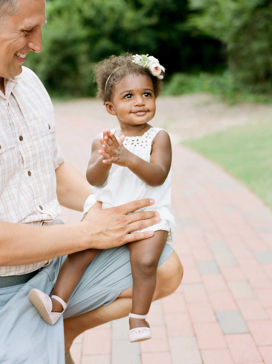 Father kneels outside smiling at adopted daughter who sits on his knee