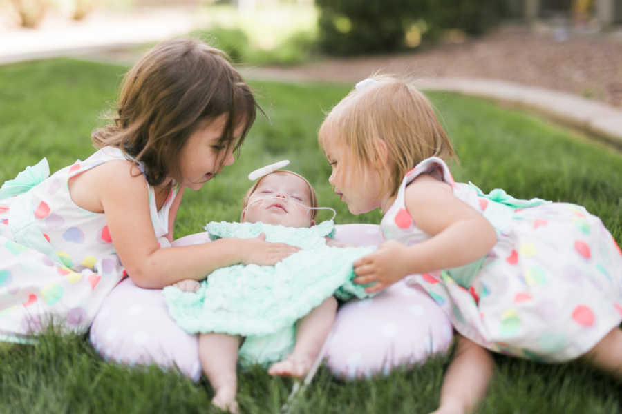 Newborn with down syndrome lays on pillow outside while her older sisters sit beside her