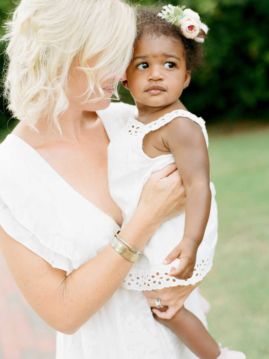 Mother stands outside in white dress holding adopted daughter who is also wearing white dress