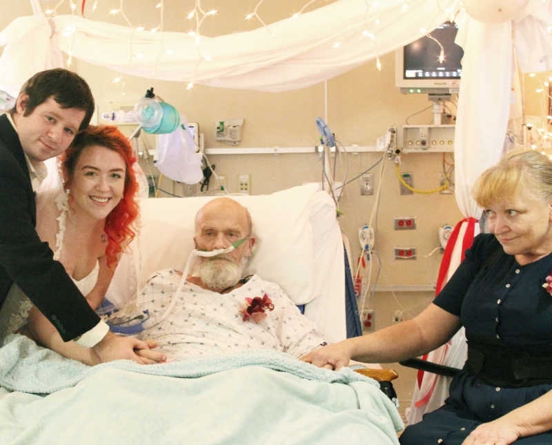 Bride stands beside groom and father with lung issues in hospital bed and her mother is on other side of bed