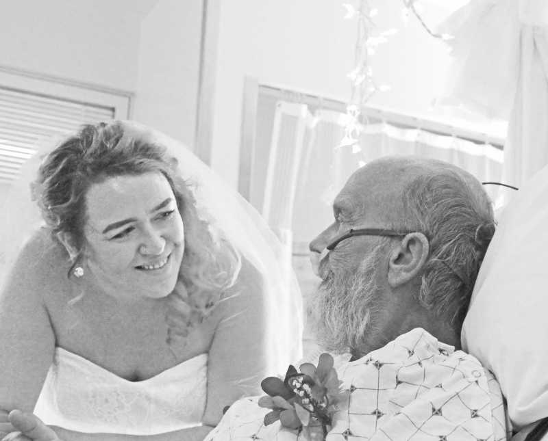 Bride leans over smiling at father who lays in hospital bed with lung issues