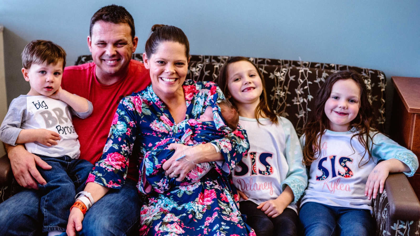 Mother smiles as she holds newborn beside two daughters and husband who holds son