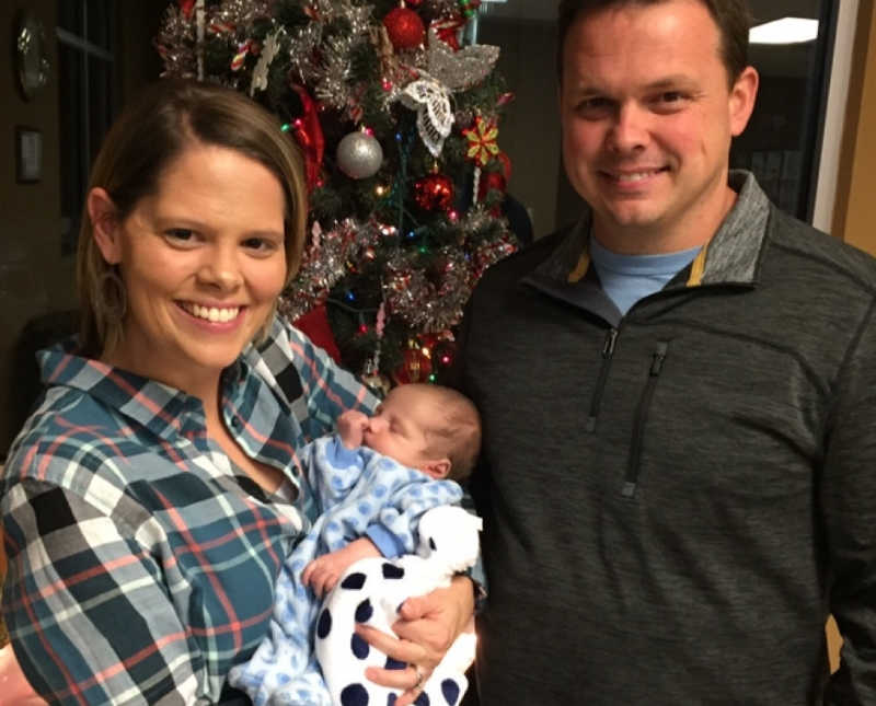 Husband and wife hold adopted newborn boy in front of Christmas tree