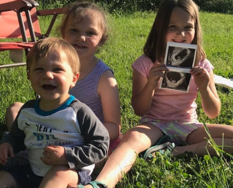 Three siblings sit on ground outside while one holds ultrasound pictures