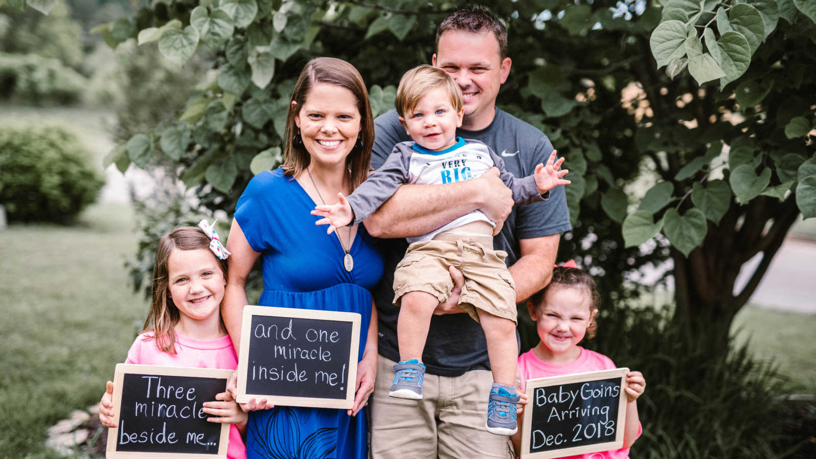 Husband smiles as he holds youngest son beside wife and two daughters who hold signs announcing new baby