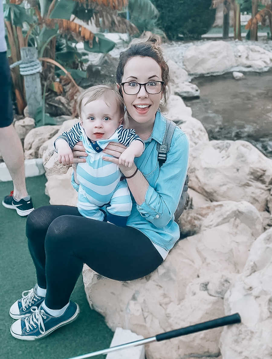 Mother sits on rock at putt putt course with son who has dwarfism on her lap