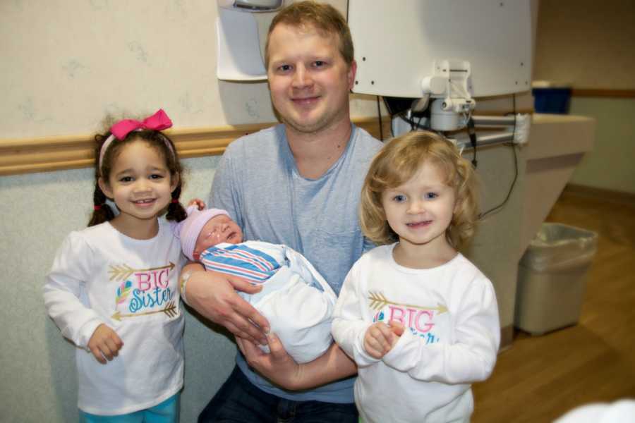 Father kneels holding newborn beside adopted daughter and biological daughter