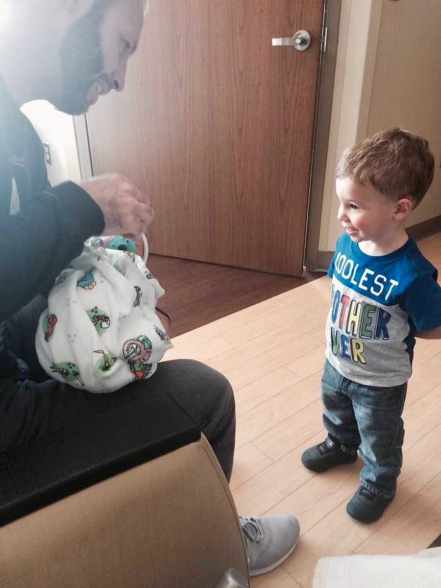 Husband sits holding newborn while first born stands in front of him staring at baby