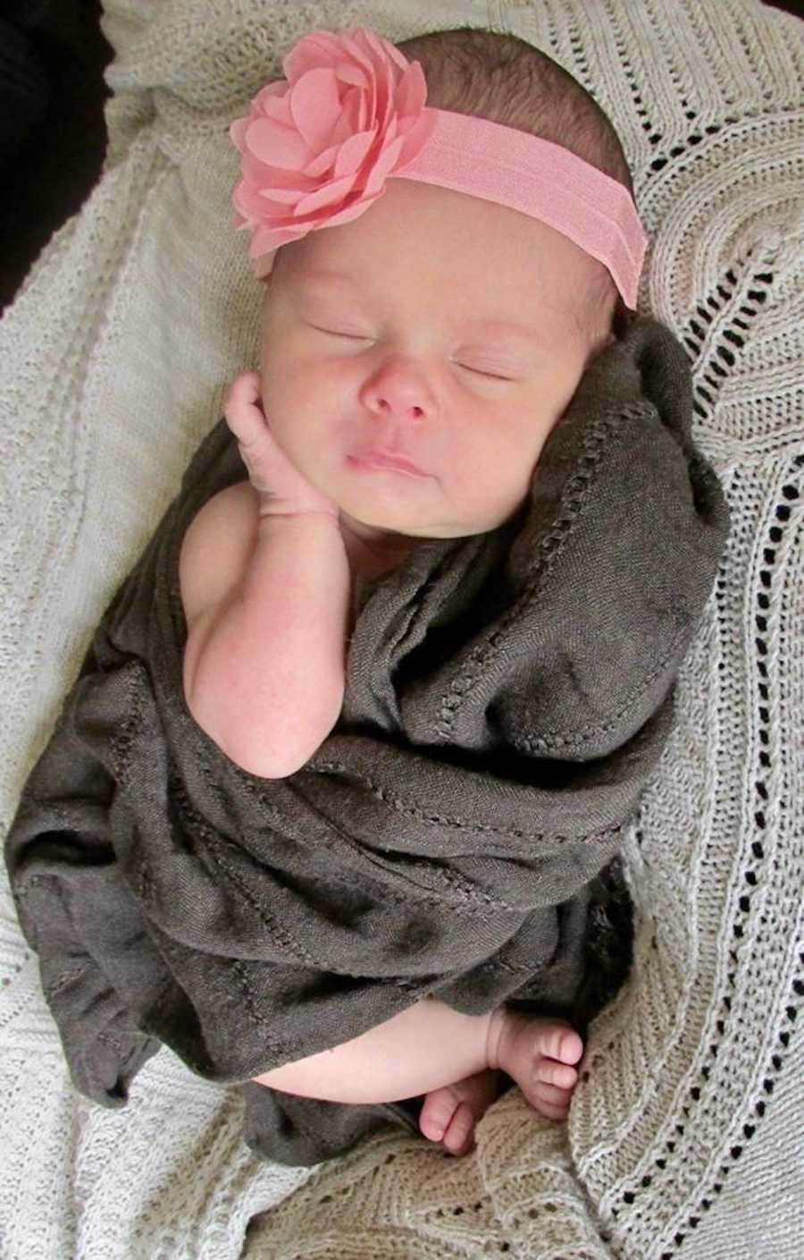 Baby girl with pink flower headband on lays sleeping swaddled in gray blanket