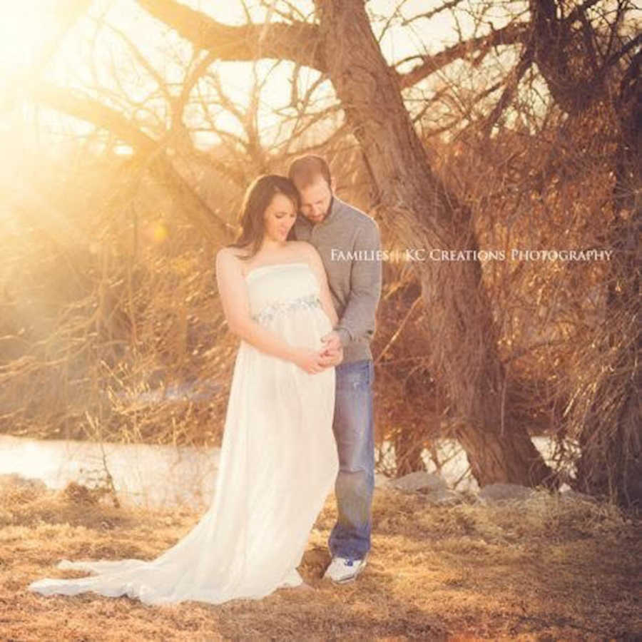 Husband stands outside beside pregnant wife in white gown as they hold her stomach